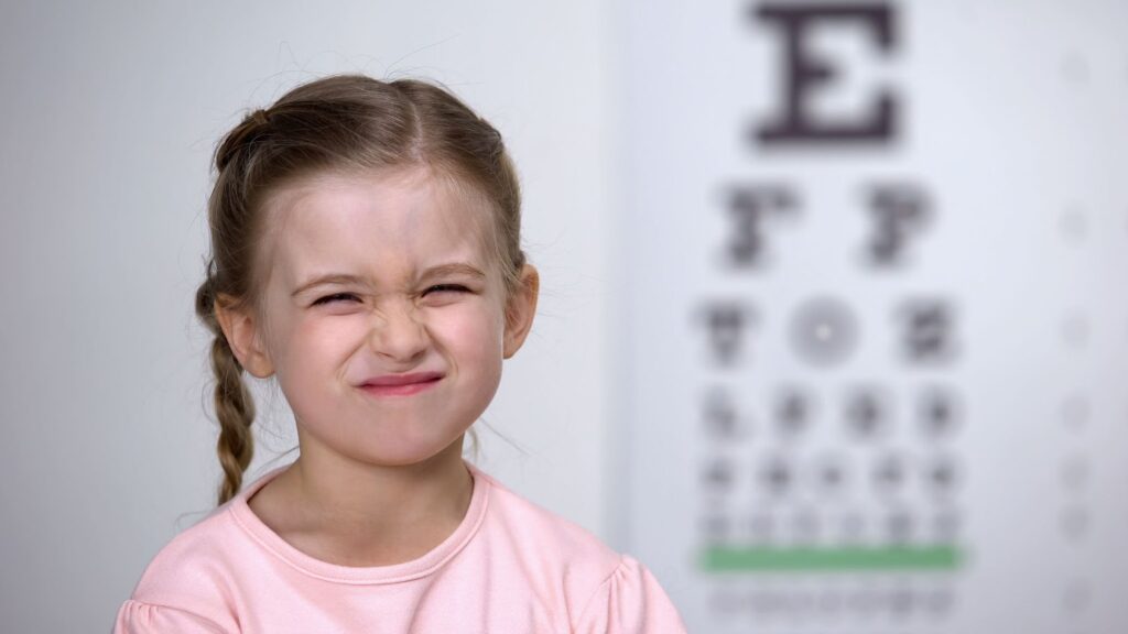 A young girl with myopia squinting her eyes with a blurry image of a Snellan chart on her right.