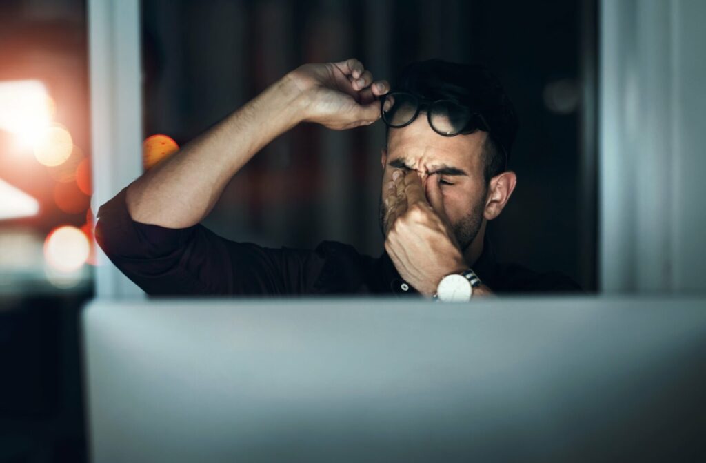 A man leans back and rubs his strained eyes while working on his computer in a dimly lit room.