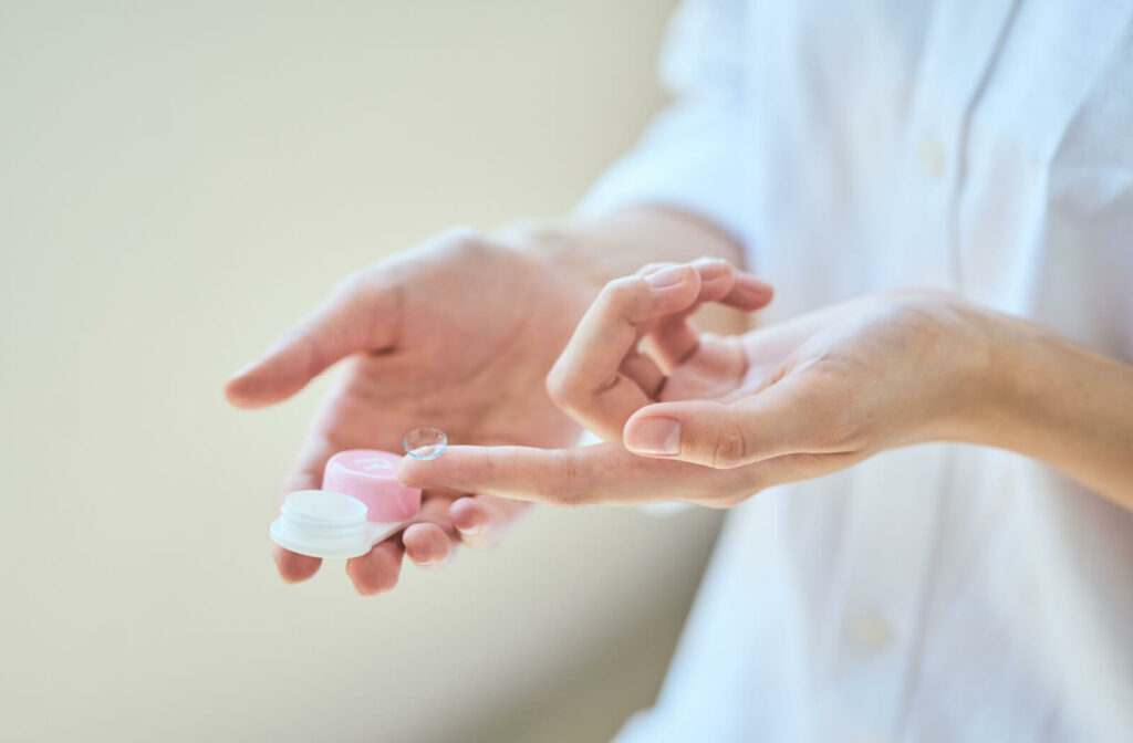 An individual in a white shirt holds a single contact lens on one finger while holding a contact lens case in the other