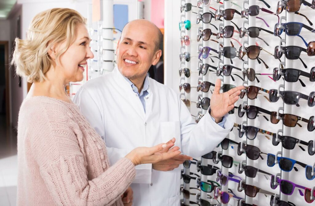 A patient with blue eyes smiling while an optometrist helps them pick out a pair of UV-blocking sunglasses from a display.