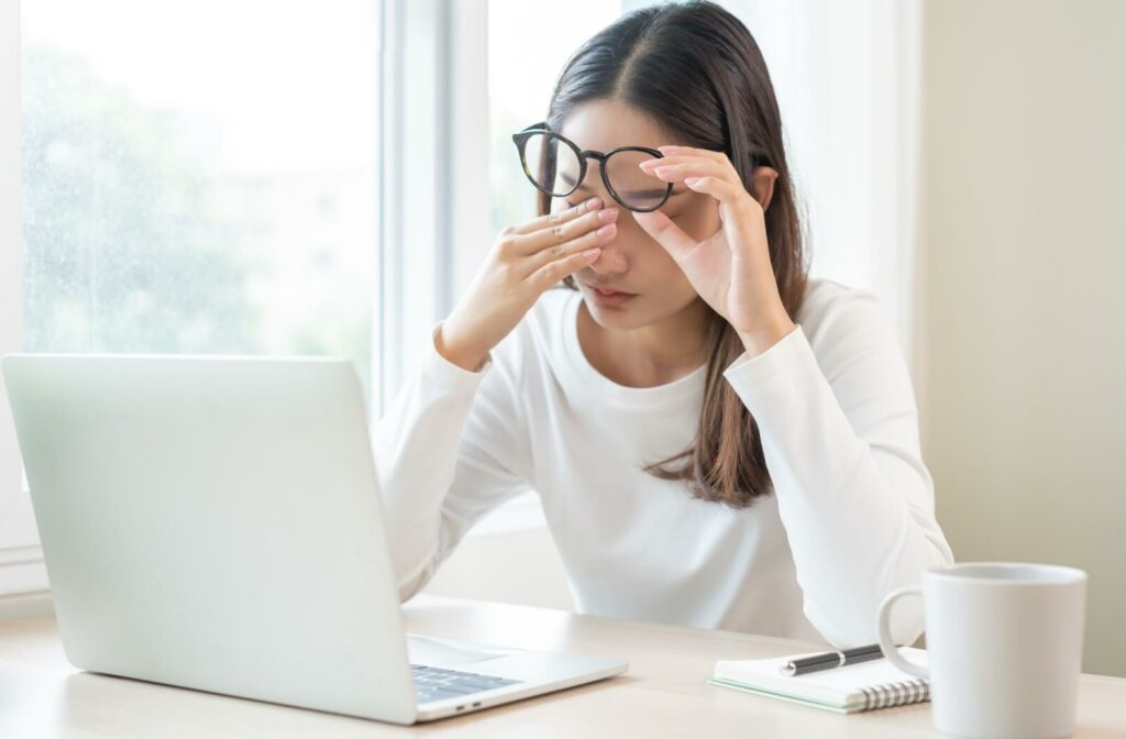Person sitting at a desk with a laptop, rubbing their eyes while wearing glasses, appearing to experience discomfort or eye strain.